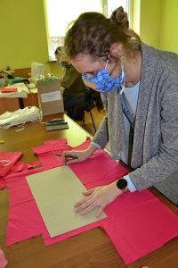 Kobieta w maseczce za pomocą tektury zaznacza na materiale wymiary. Trzyma w dłoni długopis.