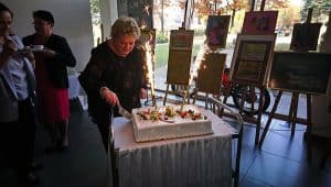 Na stole leży tort z racami. Przed nim stoi kobieta z nożem wbitym w tort. Po lewej stronie stoją osoby.