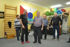 Ludzie stoją w grupce. Jest to sala gimnastyczna. Ludzie patrzą w kierunku zdjecia.