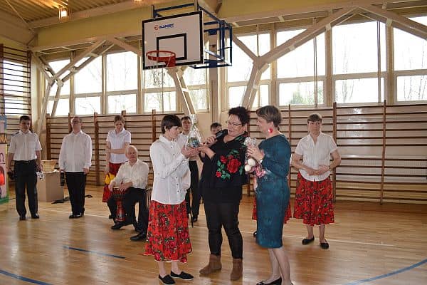 Na sali jest grupa osób . Dwie kobiety stojące obok siebie trzymają mały upominek .Obok nich stoi kobieta z podarkiem w dłoni.