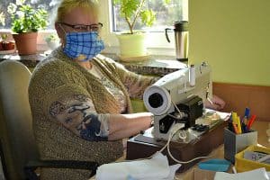 Starsza kobieta w okularach i maseczce pozuje do zdjęcia razem z maszyna do szycia.