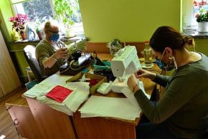 Dwie kobiety są przy stole. Jedna z nich stoi nad materiałem z długopisem. Druga kobieta siedzi trzymając kawałek materiału.