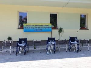 Przed budynkiem stoją naprzemiennie balkoniki z wózkami inwalidzkimi. Na wózkach leżą kule. Za nimi stoi transparent z napisem: “Warsztaty terapii Zajęciowej przy Parafii Miłosierdzia Bożego w Chełmie”.