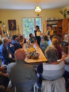 Uczestnicy siedzą przy stole zwróceni do kobiety prowadzącej warsztaty kulinarne, obok niej stoi tłumaczka PJM przodem do uczestników i obiektywu. Na stole przygotowane składniki.