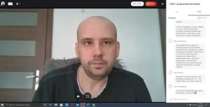 Mężczyzna siedzi na krześle w pokoju. Patrzy on w kamerę.