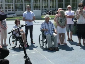 Kobieta i mężczyzna na wózkach inwalidzkich są na torze. Między nimi stoi mężczyzna. Wokół nich stoją ludzie.