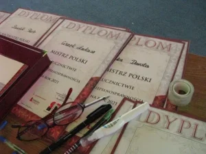 Na stole leżą dyplomy oraz okulary i długopisy.