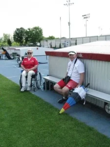 Kobieta i mężczyzna pozują do zdjęcia i są uśmiechnięci. Kobieta jest na wózku inwalidzkim. Mężczyzna siedzi na ławce.