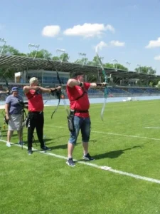 Dwóch mężczyzn stoi między biała linią i celują z łuków do celu. Obok nich stoi kobieta. Sa zwróceni bokiem do obiektywu.