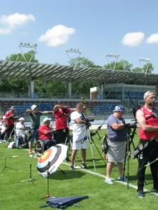 Uczestnicy stoją na linii. Część z nich celuje do celów z łuków a inni przygotowują sprzęt. Są także osoby na wózkach inwalidzkich, które strzelają. Wszyscy są zwróceni bokiem do obiektywu.