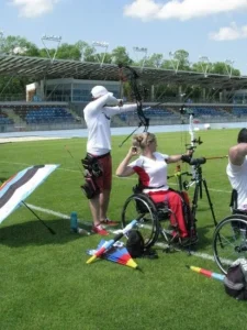 Kobieta na wózku inwalidzkim wypuściła strzałę. Osoba po jej lewej stronie celuje strzałą. Są zwrócone bokiem do obiektywu.