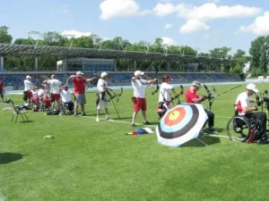 Osoby na wózkach inwalidzkich i pełnosprawne stoją razem w jednej linii i strzelają z łuków. Wszyscy są zwróceni bokiem do obiektywu.