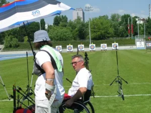 Kobieta i mężczyzna na wózku stoją na trawie. Kobieta jest zwrócona bokiem do mężczyzny oraz obiektywu, tak że nie widać twarzy.