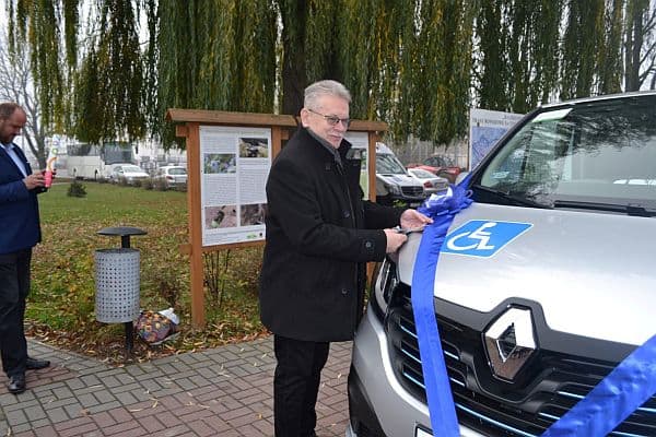 Mężczyzna stoi przed samochodem. Na samochodzie jest naklejka z osobą na wózku oraz niebieska wstążka. Mężczyzna trzyma ręką wstążkę i jest bokiem do obiektywu.