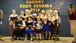 Grupa starszych kobiet jest na scenie. Trzy z nich siedzi na krzesłach a reszta jest za nimi. Mają przed sobą mikrofony na statywach.