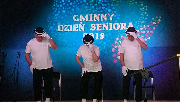 Trzech mężczyzn stoi na scenie. Sa ubrani w białe koszulki i czarne spodnie, rękawiczki białe,a na głowach kapelusze . Za nimi napis: Gminny Dzień Seniora 2019.