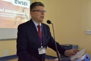 Mężczyzna w garniturze przemawia do mikrofonu zza mównicy. Za mężczyzną, na ekranie projekcyjnym wyświetlana jest grafika na której widnieje logo Wyższej Szkoły Ekonomii i Innowacji