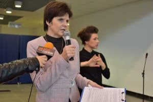 Kobieta trzymająca w jednej dłoni mikrofon a w drugiej teczkę z kartką. Po lewej stronie jest ręka z mikrofonem reporterskim wyciągnięta w stronę kobiety. W tle inna kobieta ze skrzyżowanymi ze sobą dłońmi.