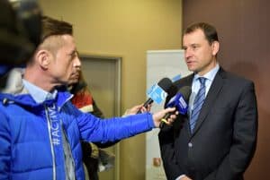 Mężczyzna trzyma w dłoni mikrofon reporterski i wyciąga go w stronę stojącego mężczyzny, który jest ubrany w wizytowy garnitur. Mężczyzna ma twarz zwróconą w stronę reportera.