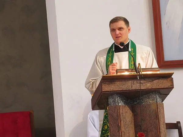 Na pierwszym planie jest młody mężczyzna w stroju księdza. Przed nim jest kazalnica i mikrofon.