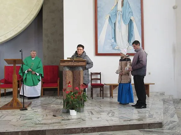 Na środku stoi kobieta przy kazalnicy i mikrofonie. Kobieta jest w kurtce. Po prawej stronie jest dwoje osób w kurtkach (mężczyzna i kobieta), a po lewej mężczyzna w stroju księdza.