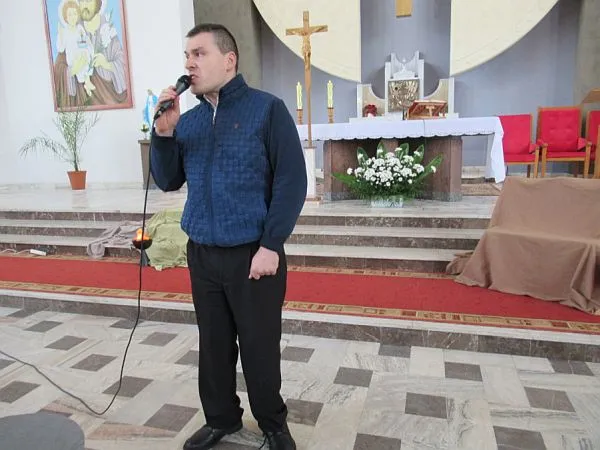 Mężczyzna stoi w kościele i trzyma w ręku mikrofon. W tle jest krzyż i ołtaż, a na ścianie obraz.