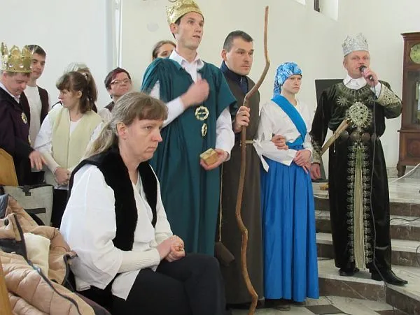 Grupa ludzi stoi. Ubrani są w różne szaty. Troje z nich ma korony na głowie. Jedna z Pań siedzi. Jeden mężczyzna trzyma w ręku długi patyk, a jeden z mężczyzn ma też mikrofon.
