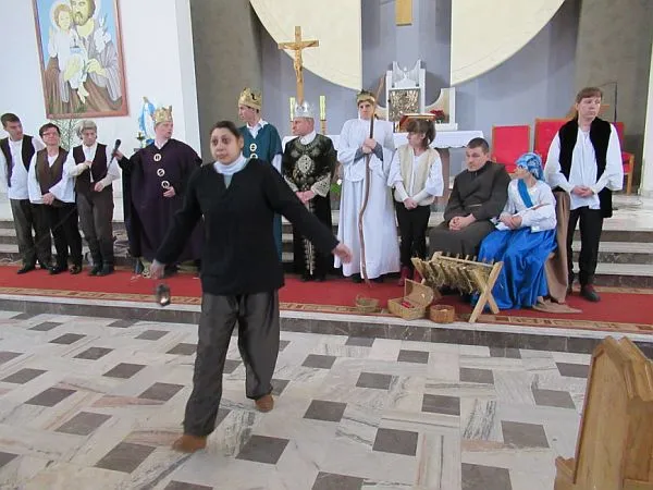 W kościele na środku stoi grupa ludzi. Przed nimi stoi jedna osoba z rozłożonymi rękami w dół. Na ścianie wisi obraz. W tle widać krzyż i ołtarz.