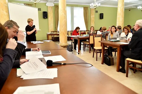 Kobieta trzymająca w dłoniach mikrofon blisko ust. Obok niej siedzą przy długim stole ludzie. Na stole leżą papiery i mikrofon. Na przeciwko stołu siedzą inni ludzie przy mniejszych stolikach.