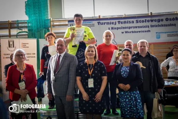 Ludzie stojący na podium. W dłoniach trzymają dyplomy i puchary. Uśmiechają się. W tle baner reklamowy z napisem “turniej tenisa stołowego”.