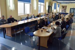 Grupa ludzi zasiadająca do dwóch równoległych względem siebie długich owalnych stołów. Na stole ludzie mają postawione filiżanki i teczki z logiem WSEI.