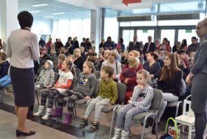 Kobieta stoi na tle siedzących ludzi na krzesłach. W pierwszym rzędzie siedzą dzieci.