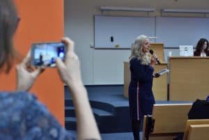 Pani o jasnym kolorze włosów i w czarnej sukience trzyma w ręku mikrofon tuż przy buzi. Druga Pani trzyma oburącz telefon, a w ekranie telefonu widać Panią trzymającą mikrofon.