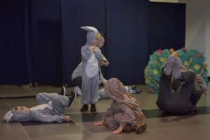 Na scenie trójka dzieci w przebraniach zwierząt. W oddali osoba wykonująca fikołka.