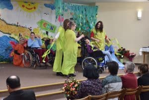 Na pierwszym planie widzimy odwróconą plecami widownię siedzącą na drewnianych krzesłach, kobieta siedząca na środku trzyma na rękach bukiet kwiatów. Na scenie widać osiem osób, trzy z nich poruszają się na wózku. W tle za jedną osobą na wózku widać kucającą kobietę – ma na sobie pomarańczową sukienkę. Za wszystkimi znajduje się kolorowa ściana z rysunkami wody, drogi, słońca i chmur oraz napis „CDN”.