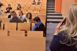 Grupa ludzi siedzi na krzesełkach w auli wykładowej. Jedna Pani stoi przed nimi i trzyma w ręku mikrofon.