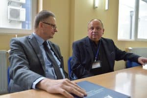 Dwóch mężczyzn siedzących przy stole. Jeden patrzy na wprost do zdjęcia, drugi patrzący w dal oraz trzymając dłoń na teczce z logiem WSEI, która jest położona na stole.