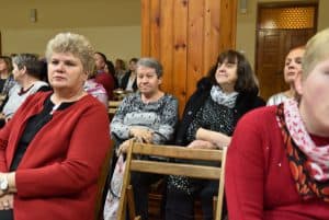 Obiektyw skierowany jest na widownię. Na pierwszym planie w środku widzimy puste krzesło, po prawej stronie widać połowę sylwetki kobiety, po lewej całą kobietę. W tle znajdują się inne osoby z widowni, drewniana kolumna oraz drewniane drzwi.