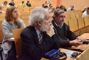 Na pierwszym tle znajduję się dwoje starszych mężczyzn siedzących na auli. Jeden z nich ma siwe włosy i ubrany jest w marynarkę, koszulę i krawat, drugi siedzący obok ma na sobie koszulę i sweter. Na drugi tle znajduje się siedząca Pani i grupa ludzi.