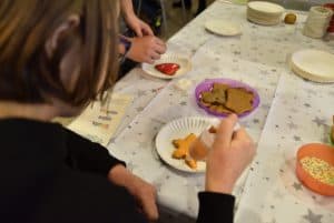 Kobieta dekoruje ciastko w kształcie gwiazdy pomarańczowym lukrem. W tle talerzyk z ciastkami i kolorową posypką dekoracyjną do ciasteczek.