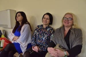 Trzy kobiety siedzą oparte o ścianę. Dwie z nich się uśmiechają.