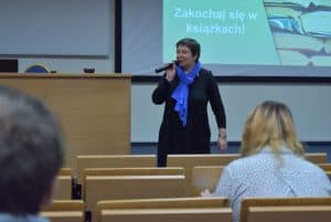 Kobieta na auli wykładowej z mikrofonem w prawej ręce, w tle na ekranie widać napis “Zakochaj się w książkach”