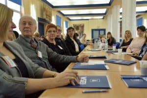 Grupa osób siedząca przy długim stole i patrząca przed siebie, na wprost do zdjęcia bądź na siebie nawzajem.