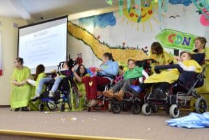 Na scenie widać 9 osób, cztery z nich to osoby z niepełnosprawnością na wózku – znajdują się one na pierwszym planie. W tle po lewej stronie widać ekran do rzutnika oraz wyświetlane na nim „Witamy. 30 lecie Dziennego Ośrodka Adaptacyjnego dla Dzieci i Młodzieży z Niepełnosprawnością Intelektualną”. Za wszystkimi znajduje się kolorowa ściana z rysunkami przestawiającymi wodę, drogę, słońce i chmury oraz napis „CDN”.