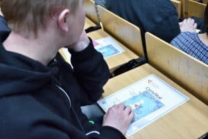 Mężczyzna siedzi przy stoliku na auli. Prawą dłoń ma opartą na stoliku na kartach, a lewą ręką podpiera sobie głowę o ławkę.