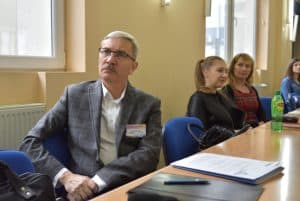 Na pierwszym planie zdjęcia znajduje się mężczyzna, w podeszłym wieku, który siedzi przy stole. Patrzy na wprost do zdjęcia. Ręce ma założone na siebie. W oddali trzy kobiety siedzące przy tym samym stole.