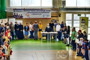 Ludzie stojący na sali gimnastycznej. Na drabinkach gimnastycznych wisi baner “Turniej tenisa stołowego osób z niepełnosprawnością intelektualną”