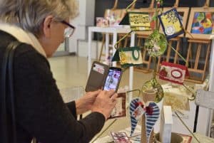 Kobieta robiąca smartfonem zdjęcie rękodzieła o kształcie figury choinki z przywieszonymi do niej kolorowymi kartkami.