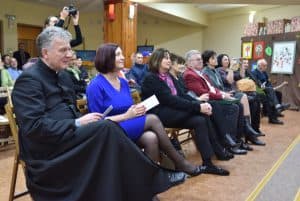 Obiektyw skierowany jest na widownię. W pierwszym rzędzie siedzi dziewięć osób – pięć kobiet i czterech mężczyzn, najbardziej po lewej znajduje się ksiądz w sutannie. W tle znajdują się inne osoby, jedna z nich używa aparatu fotograficznego.