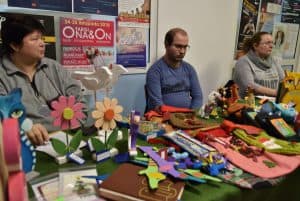 Troje ludzi siedzi przy stole na którym znajdują się rękodzieła. Dwie kobiety i mężczyzna. Wśród rękodzieł znajdują się między innymi kwiatki i ptaszki.